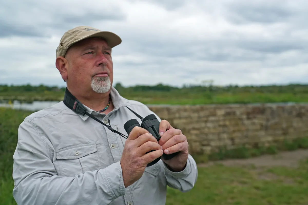 Older birder in a cap with binoculars on a meadow