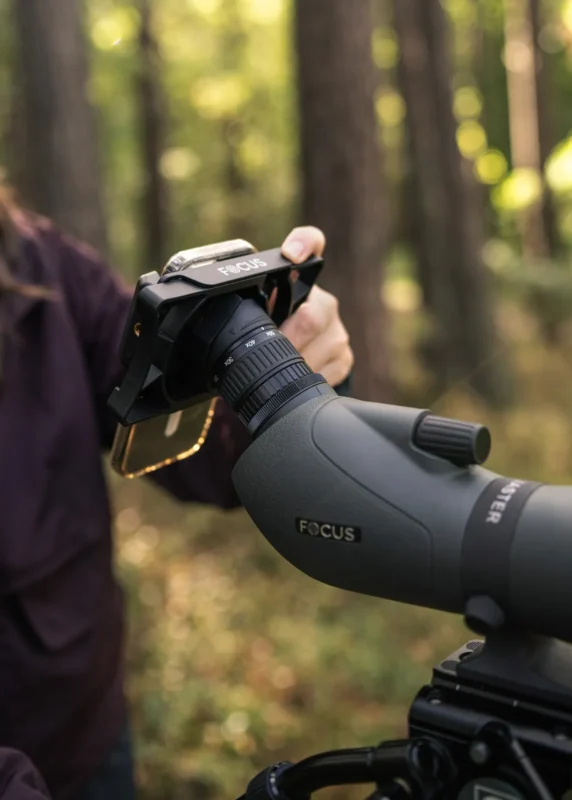A hand fitting a phone adapter with a smartphone onto a spotting scope eyepiece