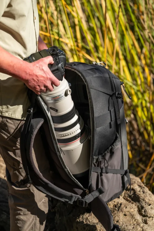 A hand lifts a camera with a large telephoto lens out of an open camera backpack