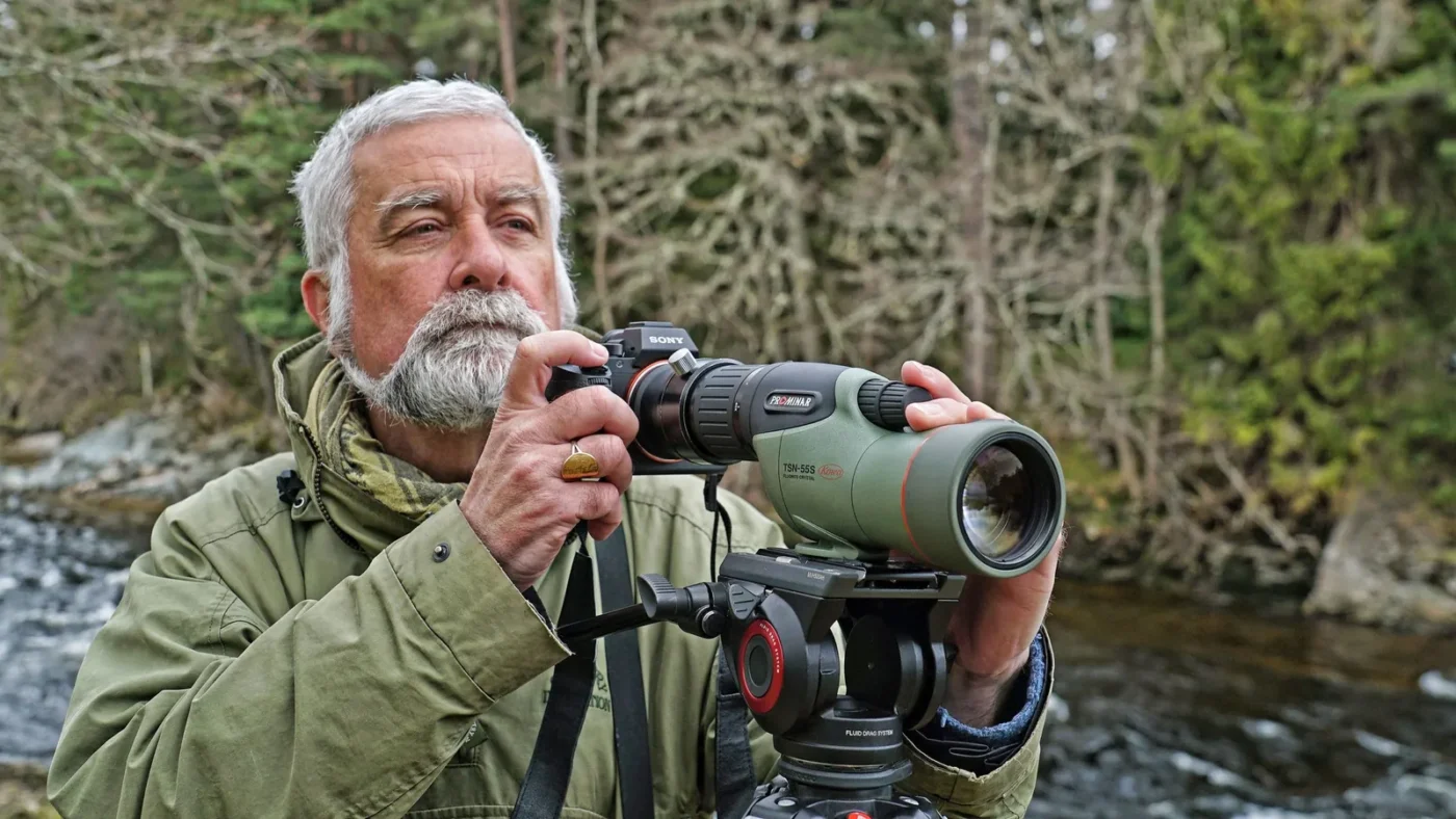 Grey-bearded man looking through a spotting scope by the water