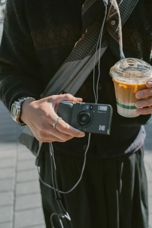 Hand holding a compact black Kodak film camera, an iced coffee in the other hand