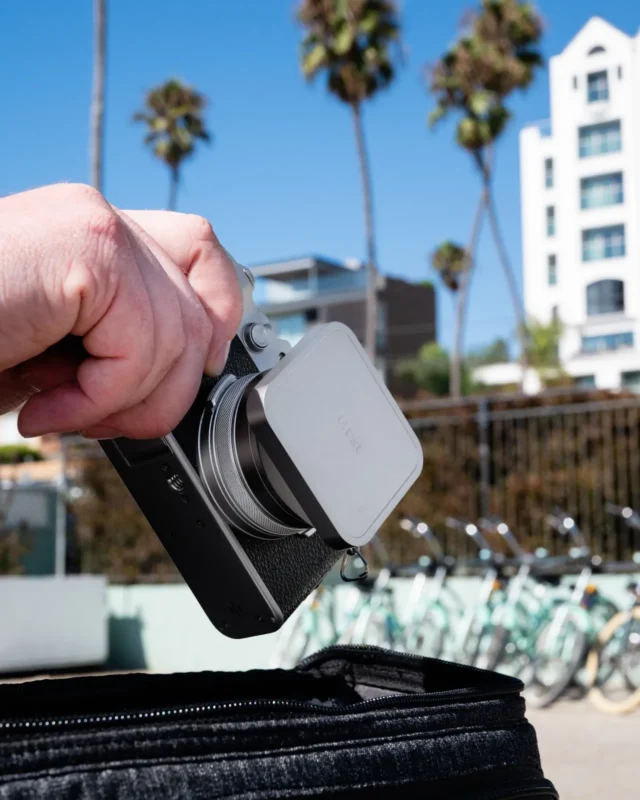 Hand holding a compact camera with a lens cover on a street