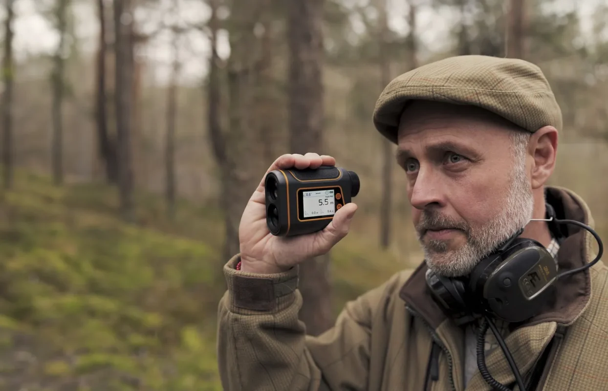 Hunter in a flat cap reading a rangefinder in the forest