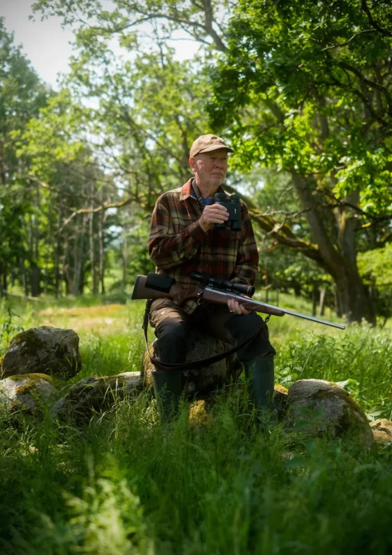 Hunter sitting in a green glade with a rifle and binoculars