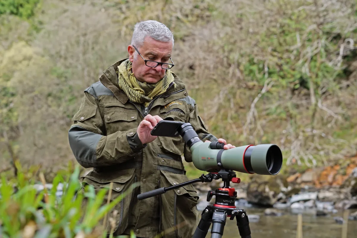 Man framing a shot on a phone attached to a tripod-mounted spotting scope