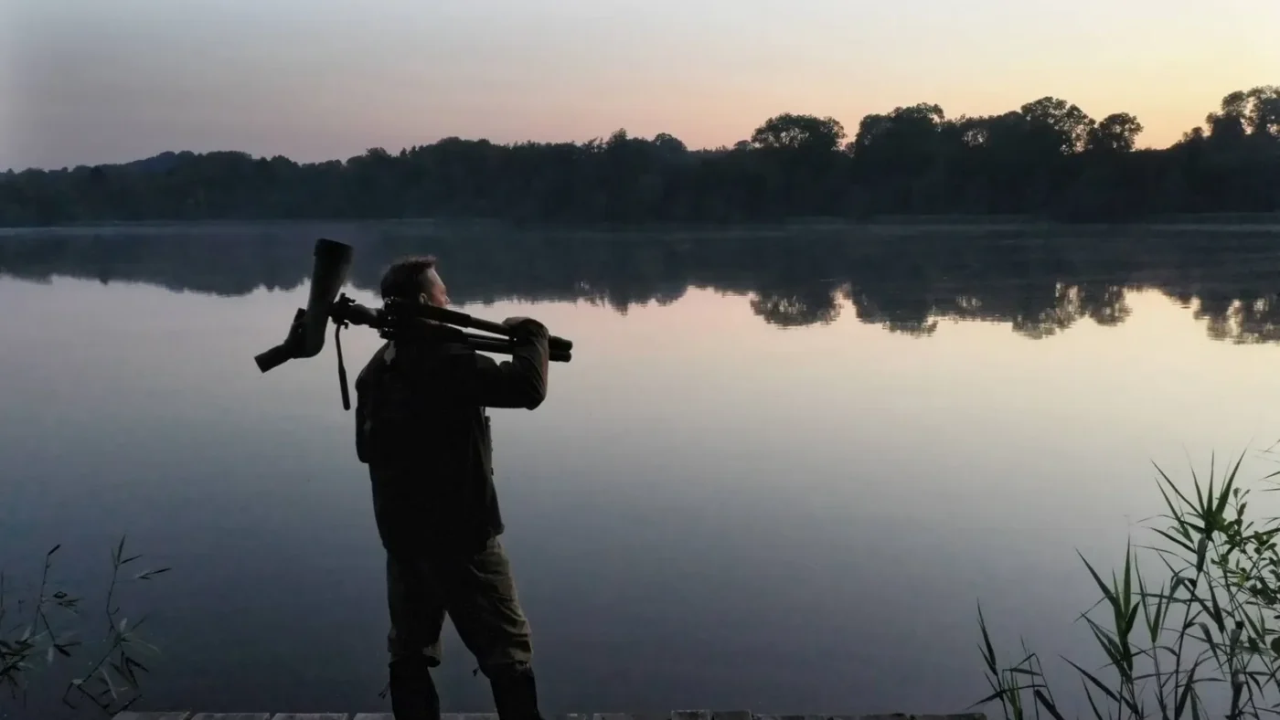 Silhouette of a person at a spotting scope by a still lake at dusk