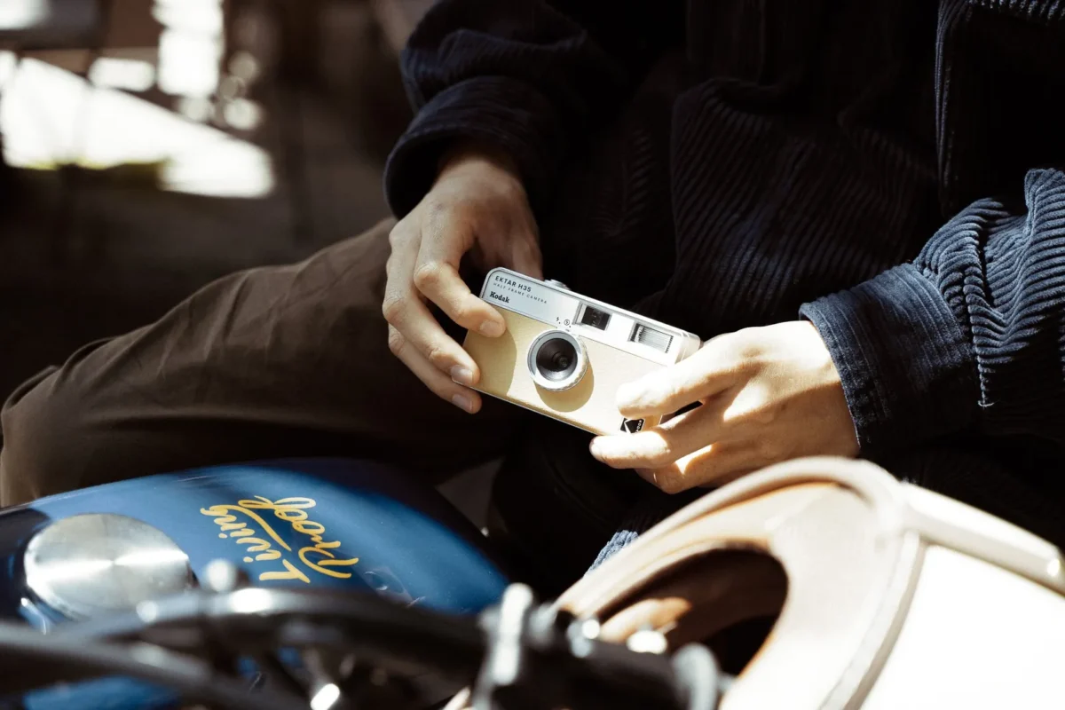 Two hands holding a beige compact film camera with fingers clear of the lens, a motorcycle in the foreground